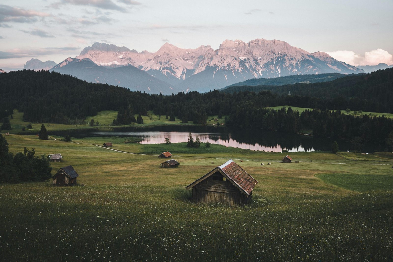 Explore the serene Bavarian landscape in Krün, Germany, with lush fields, a tranquil lake, and majestic mountains at dawn.
