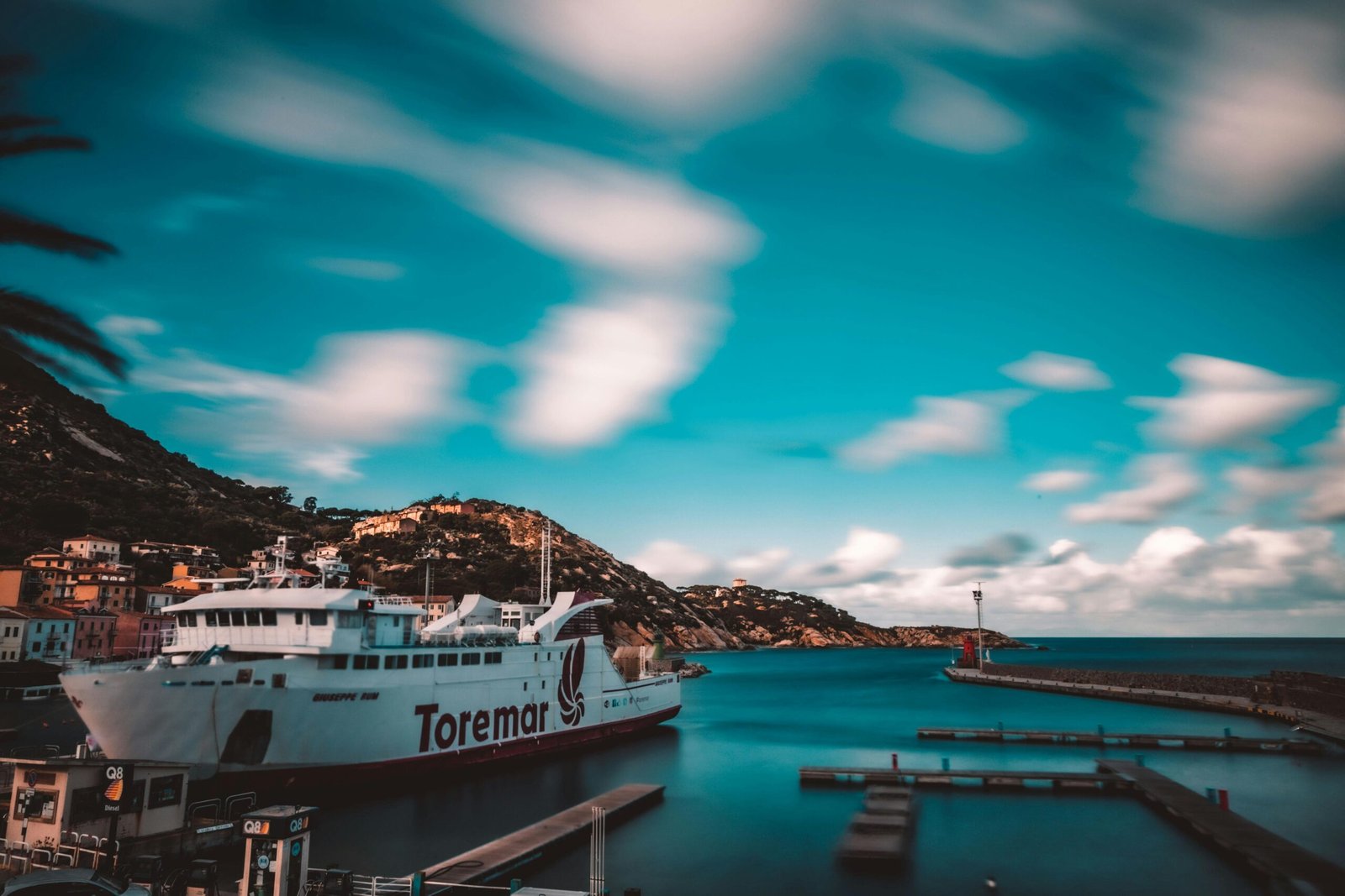 Vibrant Toremar ferry docked at a serene Tuscan port, surrounded by hills and blue sea.